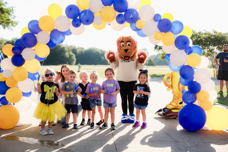 Young students smile for a group photo outdoors during a school event with their school's lion mascot