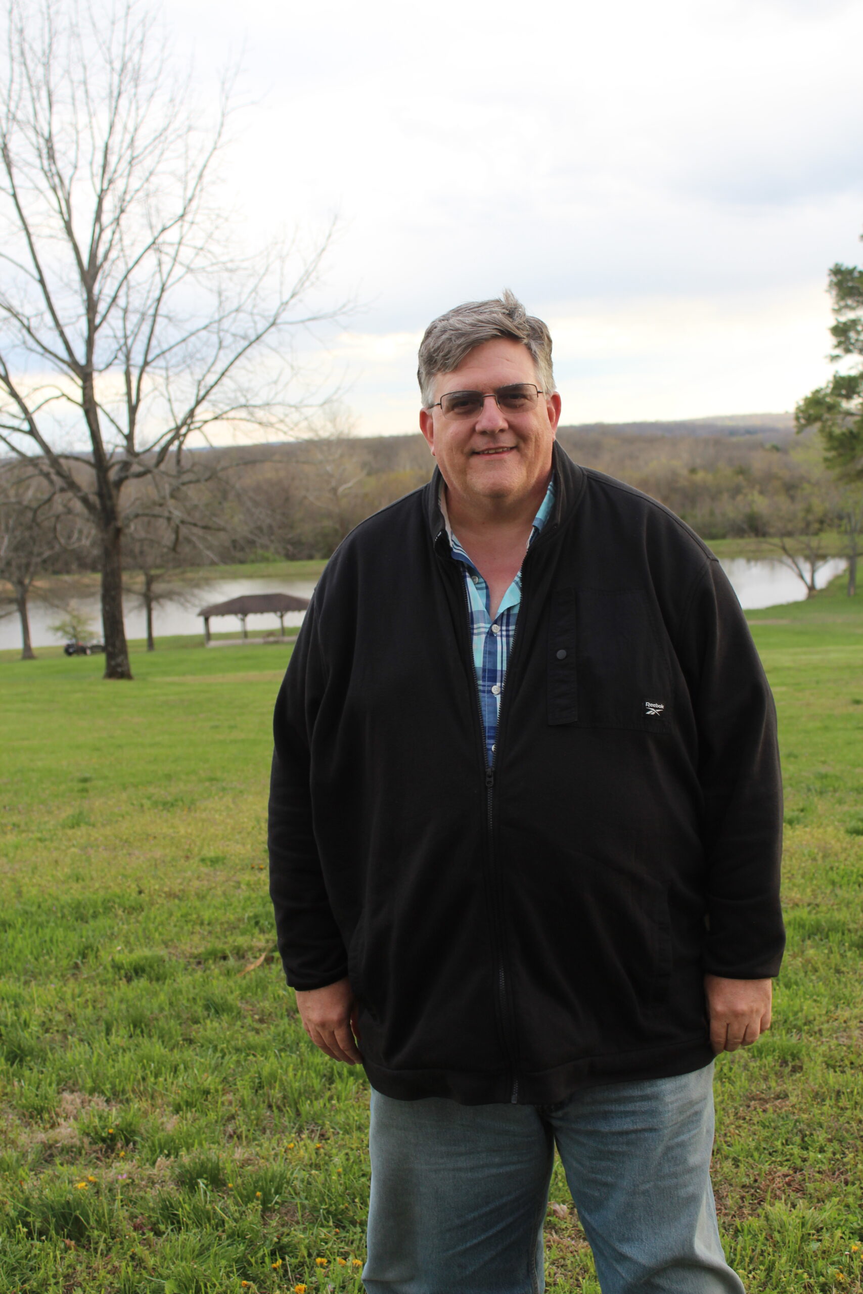 Man in black jacket stands in grassy field near lake. Outdoor portrait.