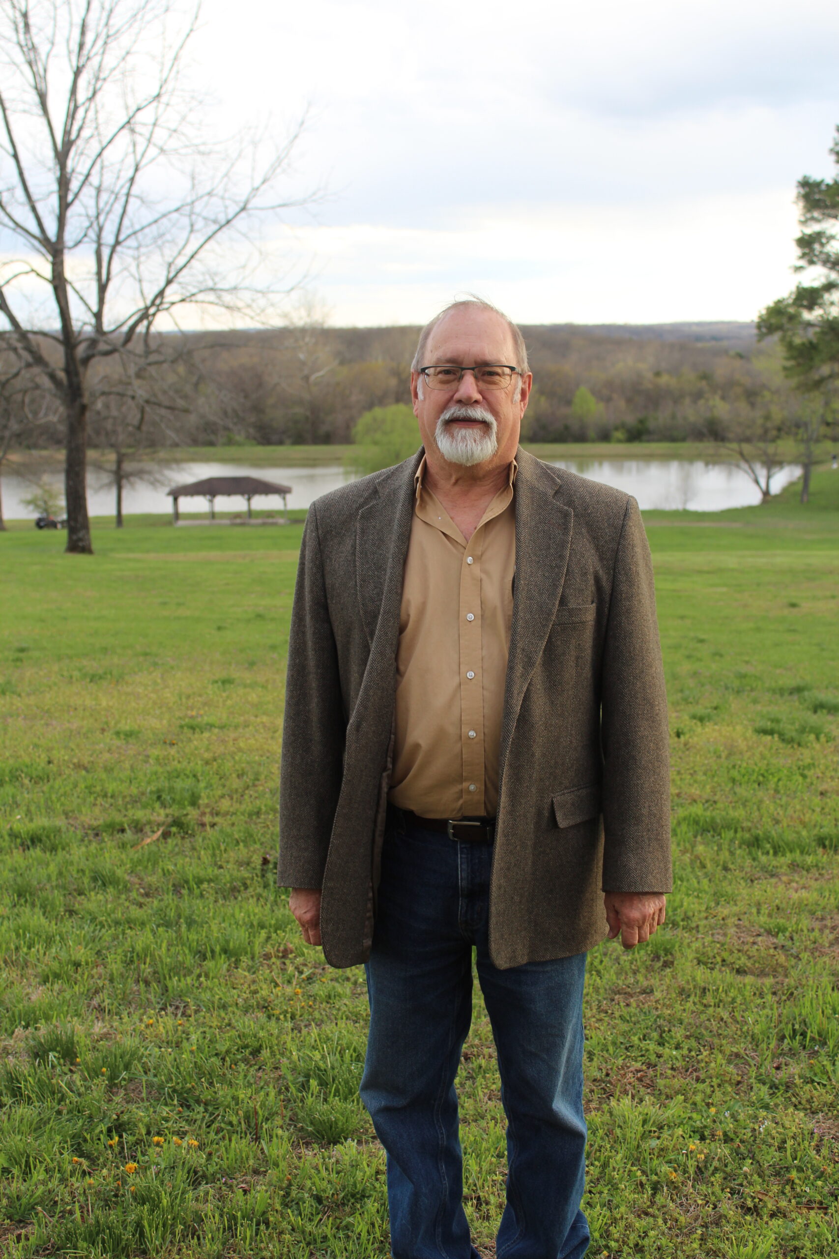 Man in blazer stands outdoors near lake. Staff photo.