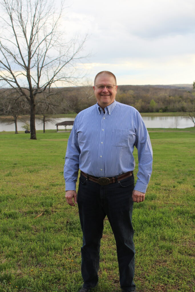 Man in blue striped shirt standing outside near a lake and trees.