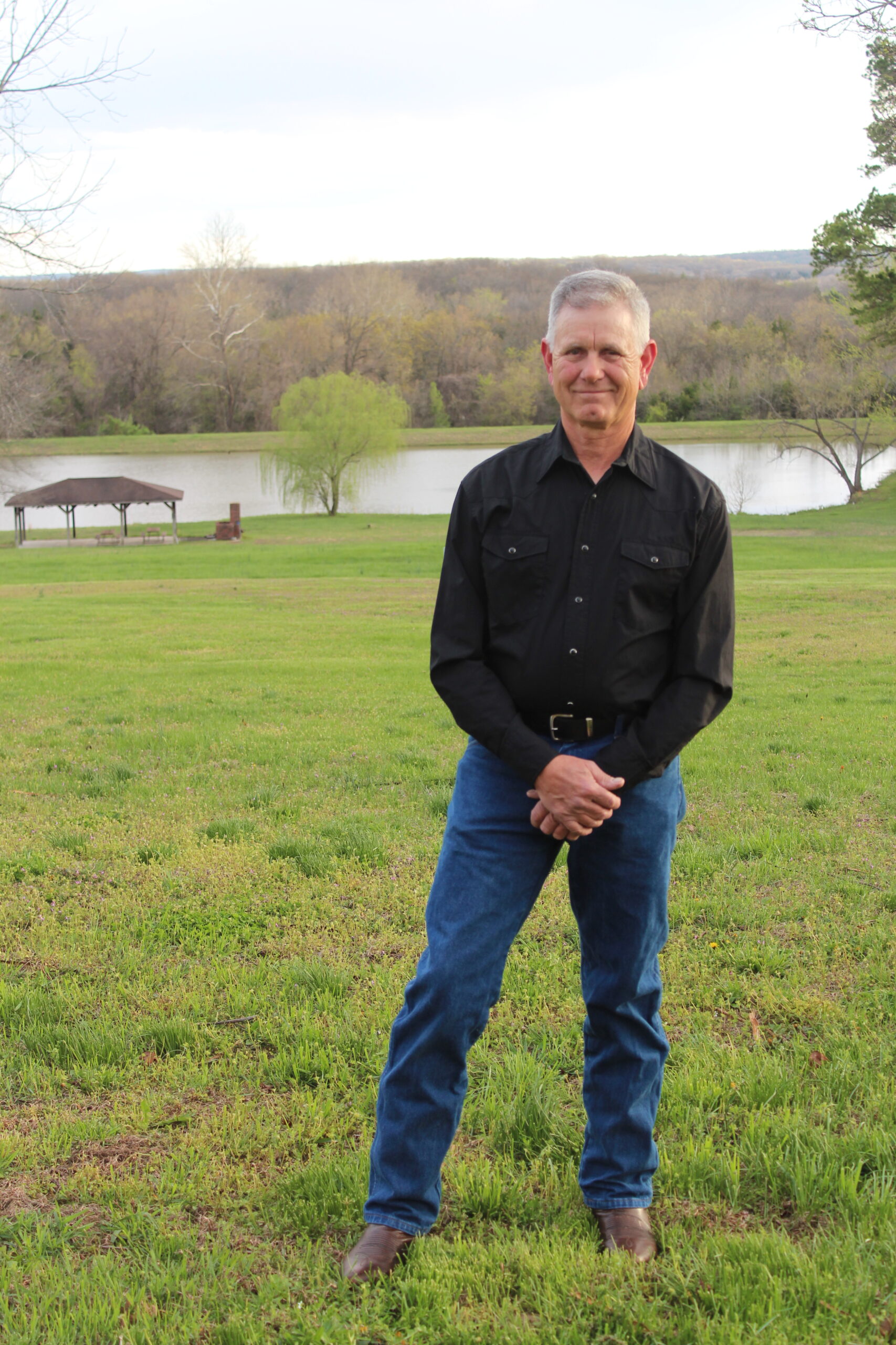 Man in black shirt and jeans stands on green lawn near lake.