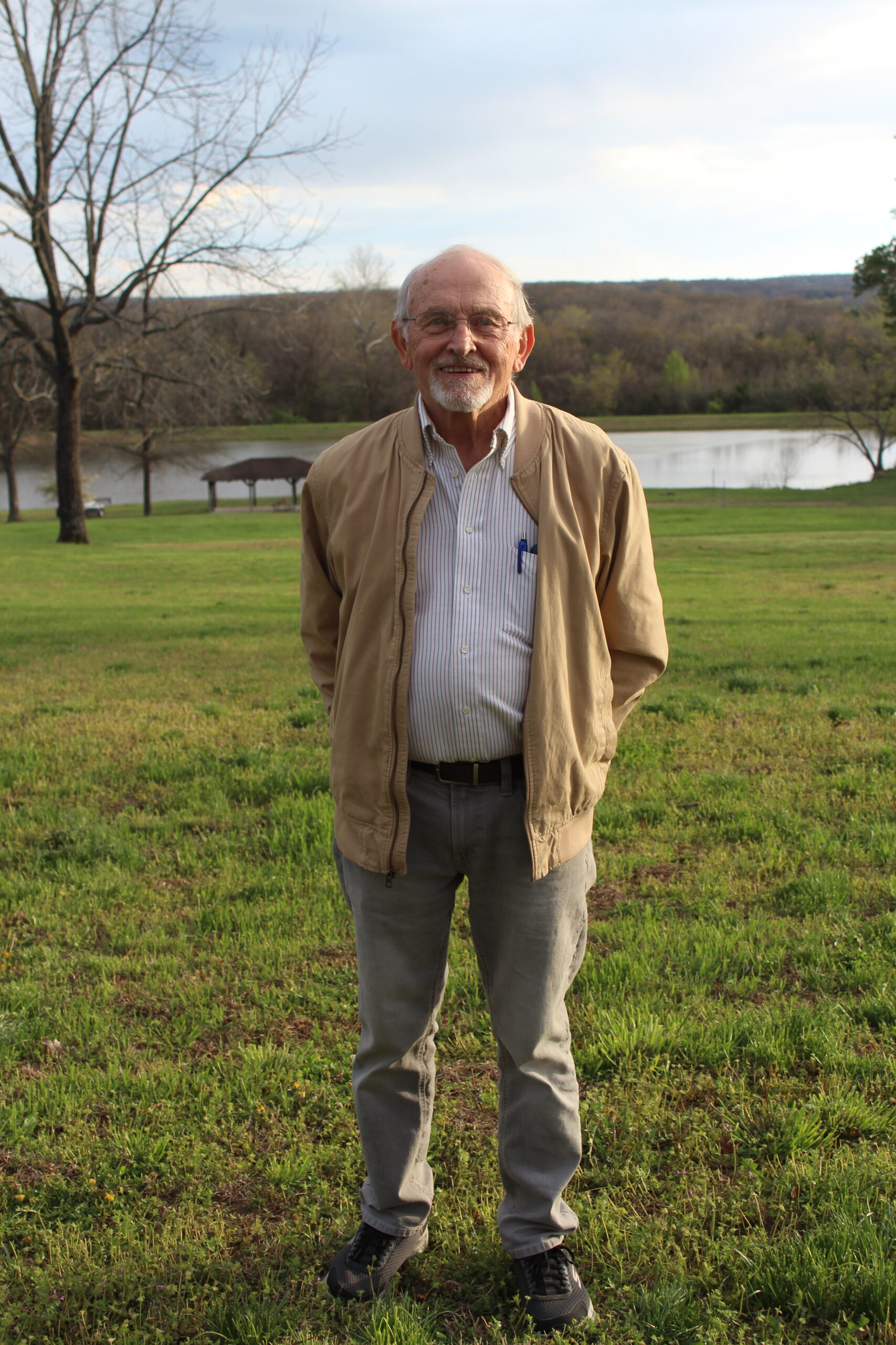 Smiling senior man in park setting with lake, trees, and green grass.
