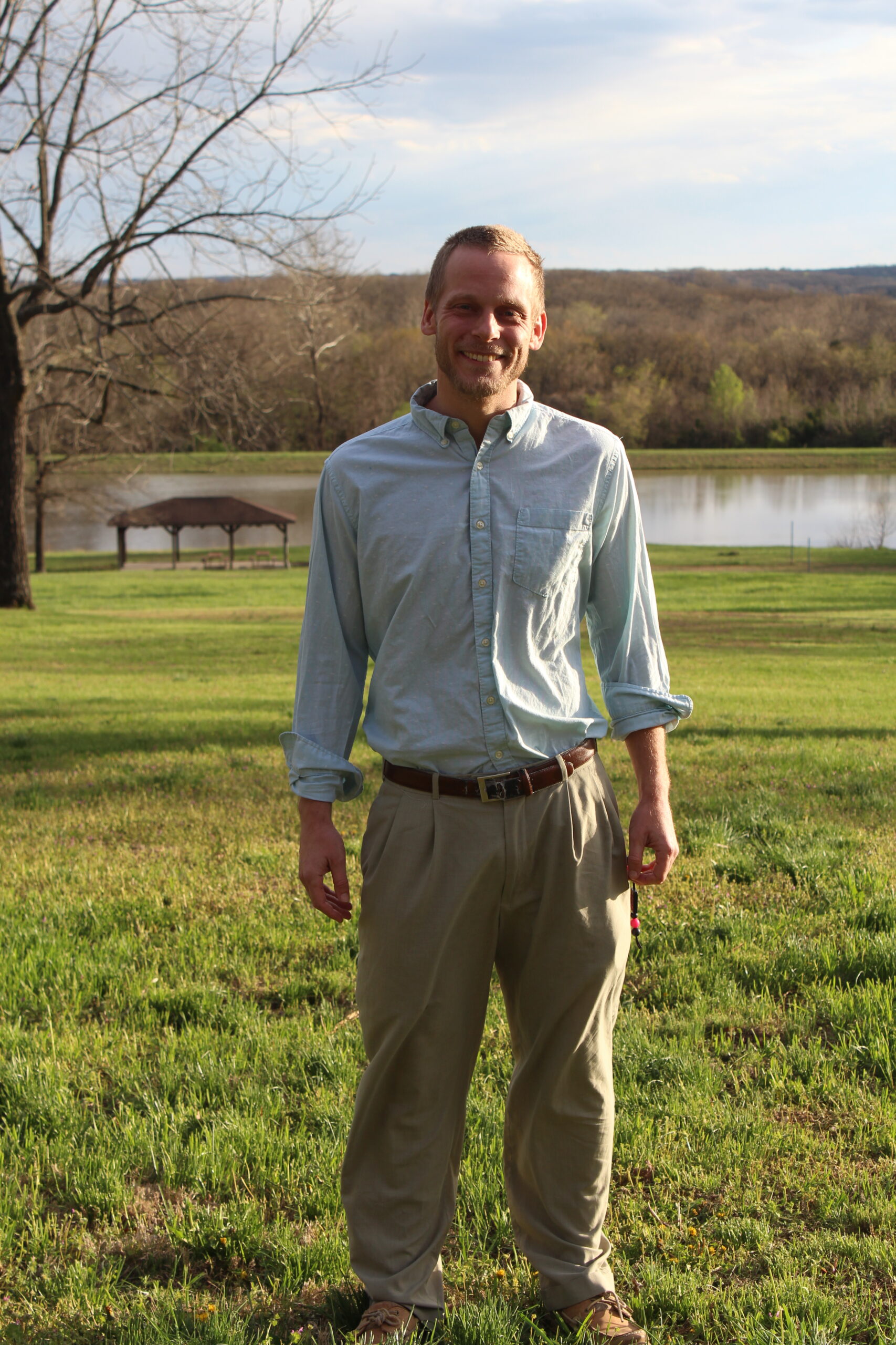 Man in light blue shirt and khakis stands smiling in a grassy field near a lake.