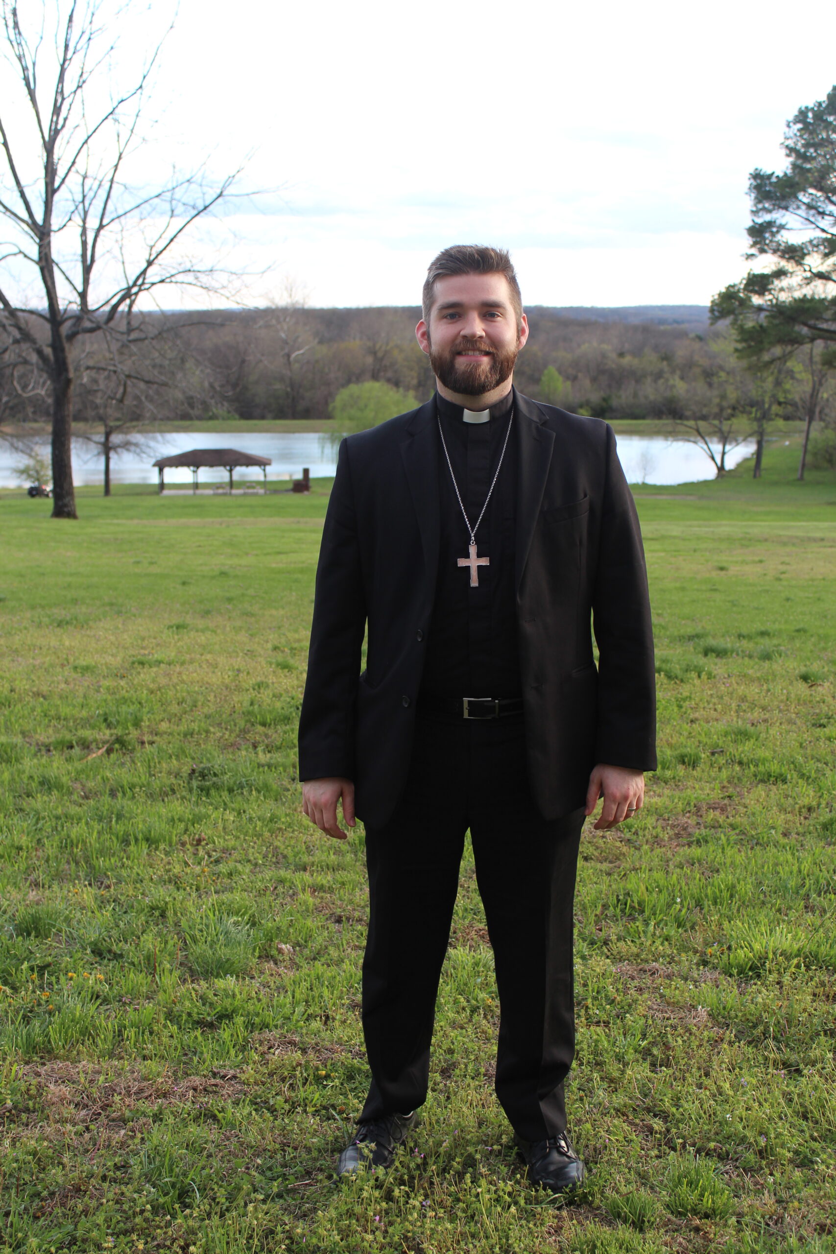 Man in clerical collar and black suit standing in a grassy field.