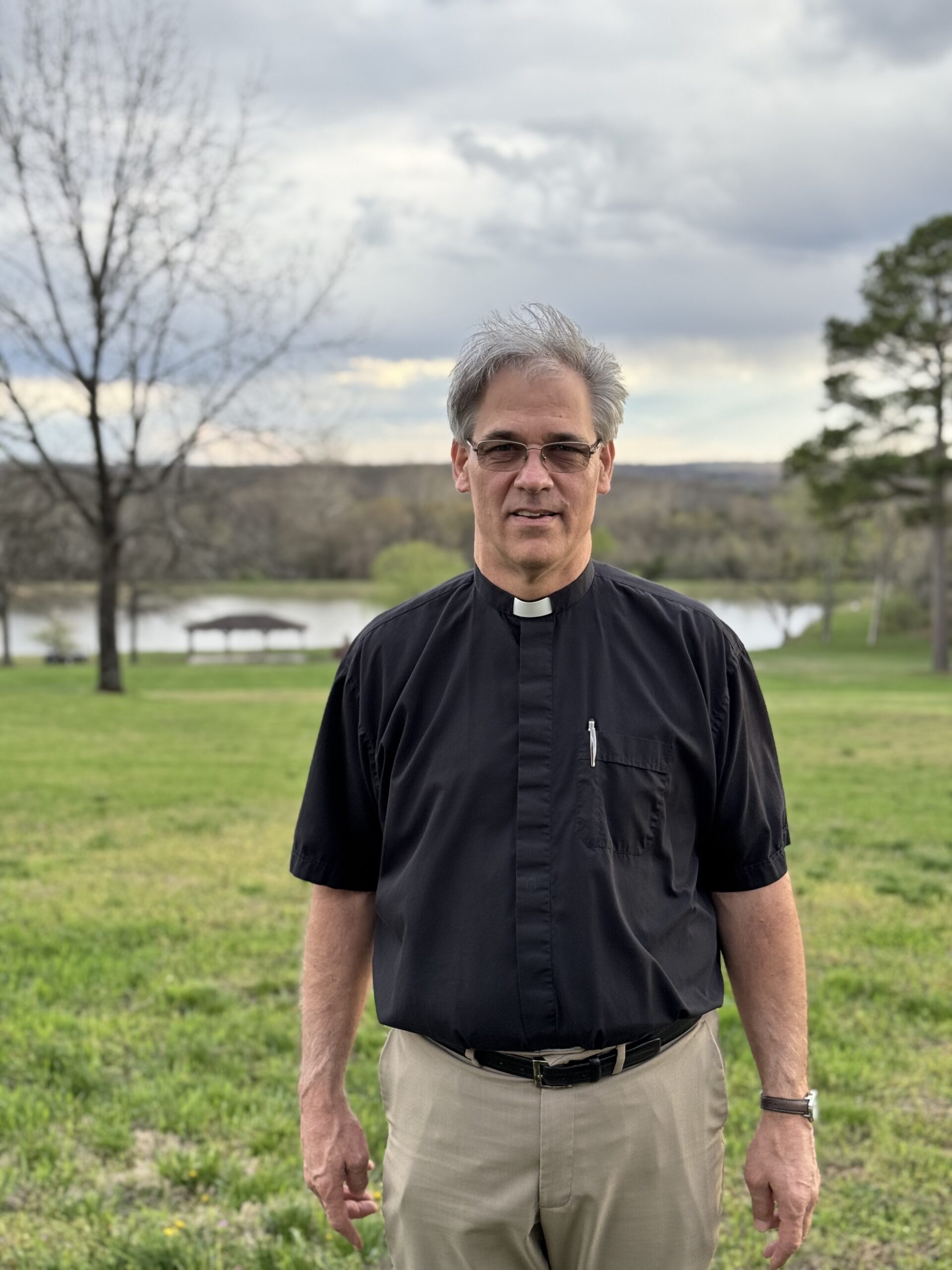 Staff photo of a priest in a clerical collar standing outdoors on a grassy field.