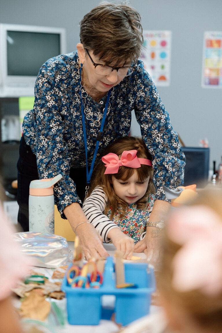 Teacher helping a young student with a craft project at a table. Learning and tuition assistance.