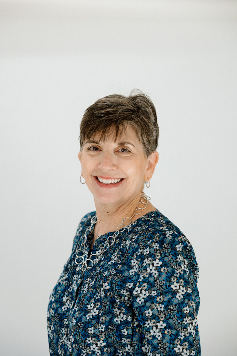 Portrait of a smiling woman with short hair wearing a blue floral blouse and silver necklace.