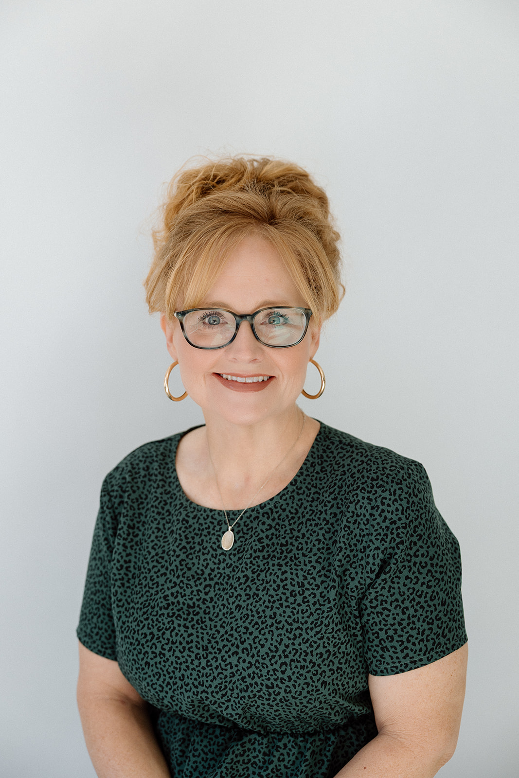 Professional woman with red hair and glasses smiles in a green patterned top.
