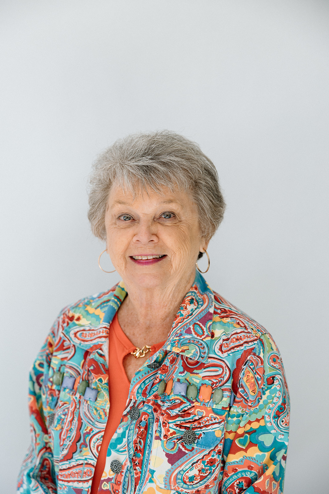 Portrait of a senior woman with short gray hair, wearing a colorful paisley jacket and orange top.