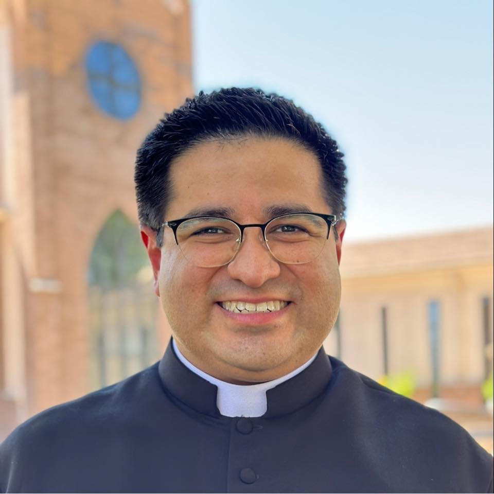 Portrait of Fr. Daniel Cruz smiling in his clerical collar, outdoors.