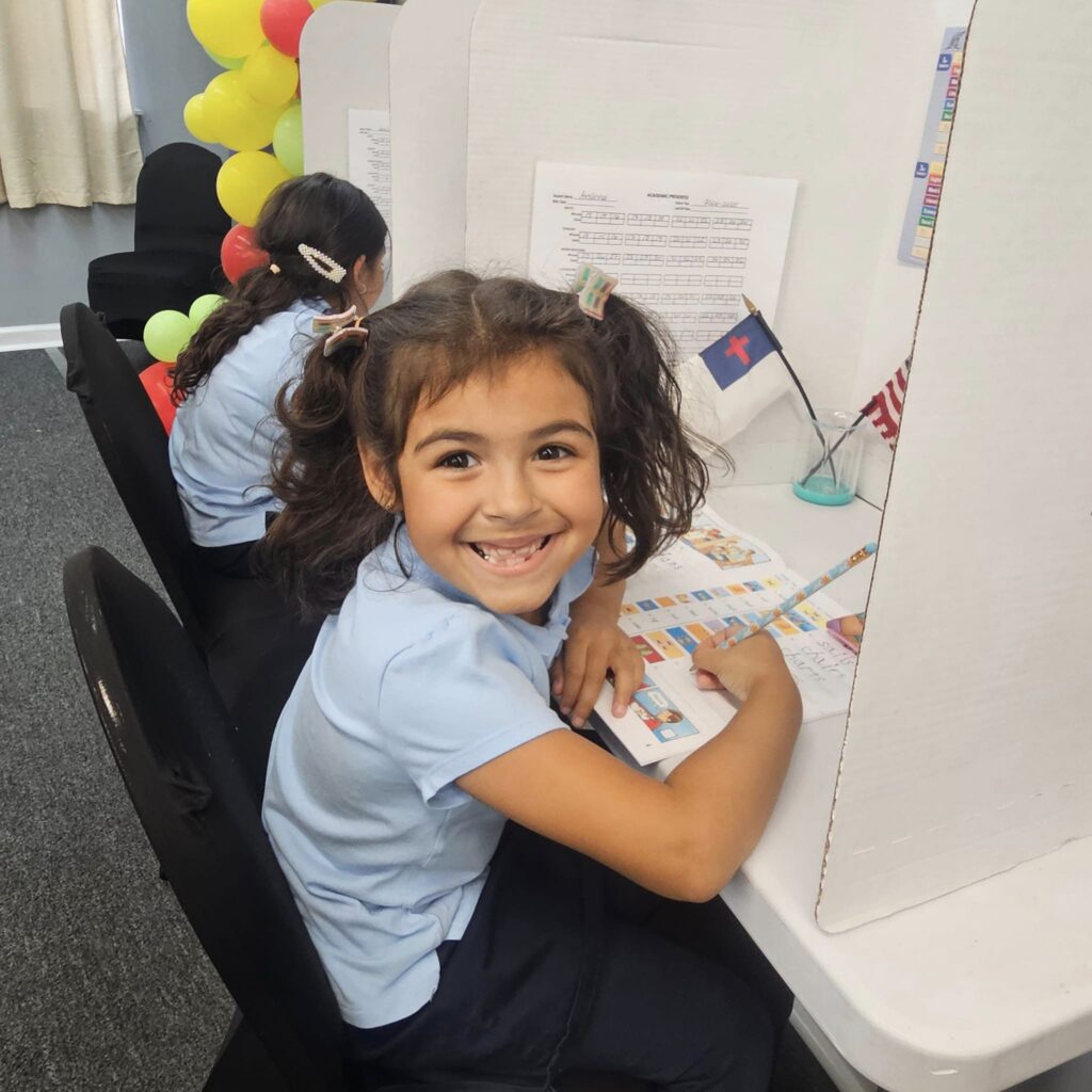 Girl in blue shirt smiles while working at her desk in a classroom setting.