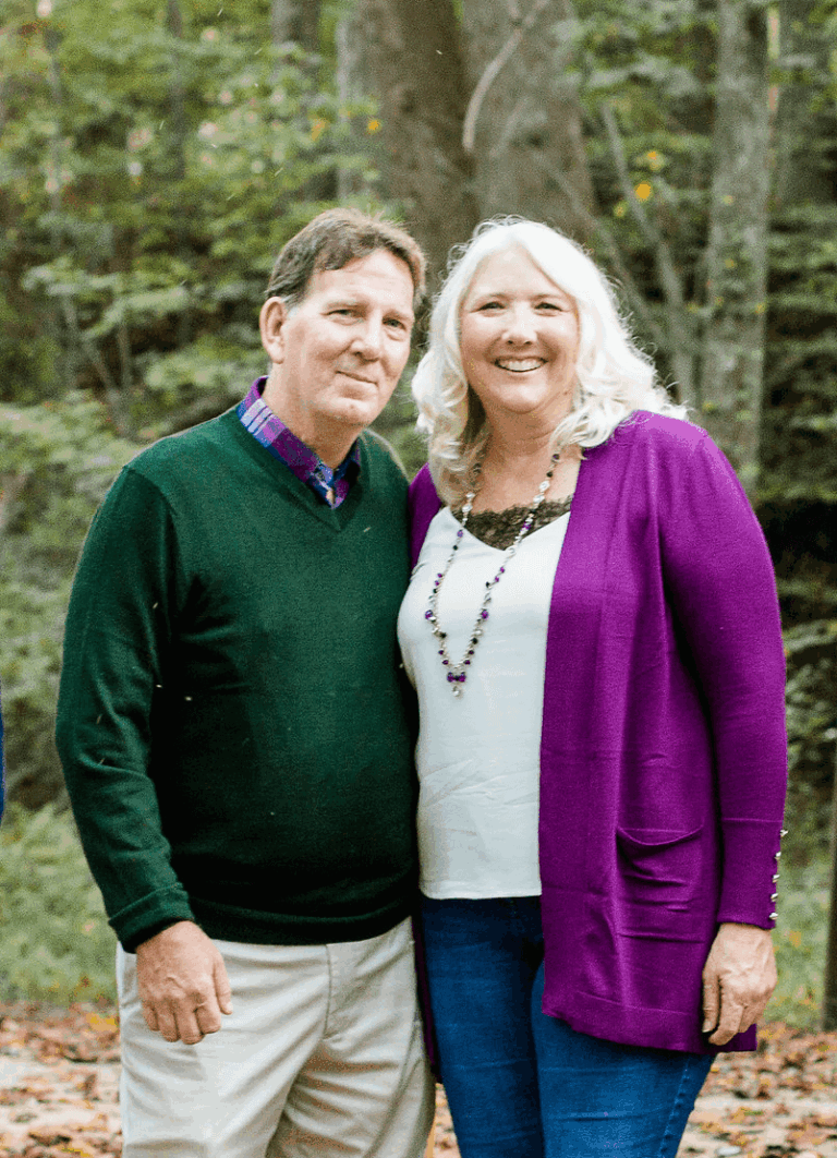 Couple smiling outdoors. Man in green sweater, woman in purple cardigan.