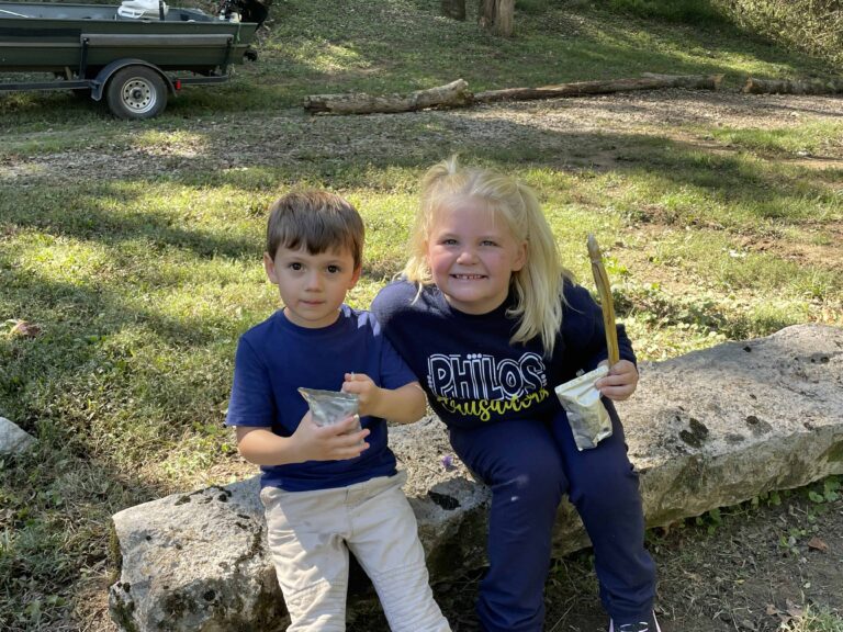 Two smiling kids sit on a stone, holding snacks outdoors.