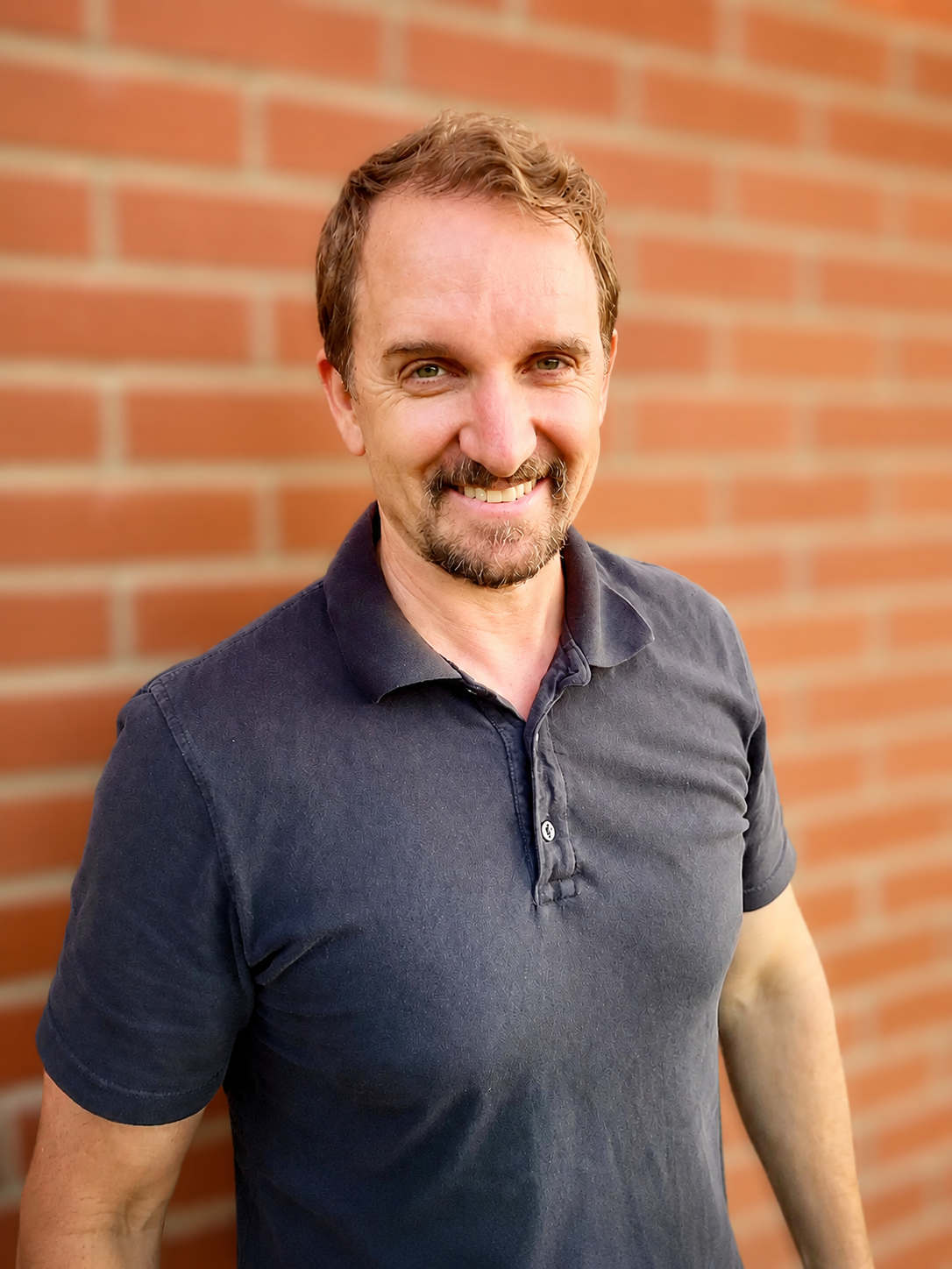 Stuart Sloan, smiling man with goatee against brick wall