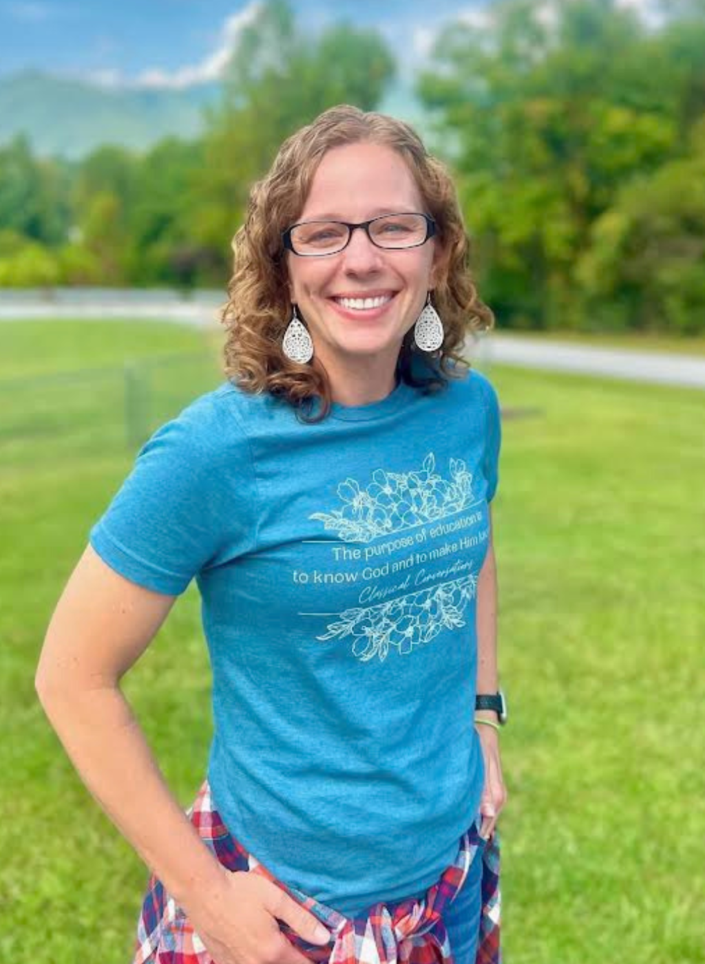 Tiffany Berg smiles in a blue t-shirt with text about education and Classical Conversations.