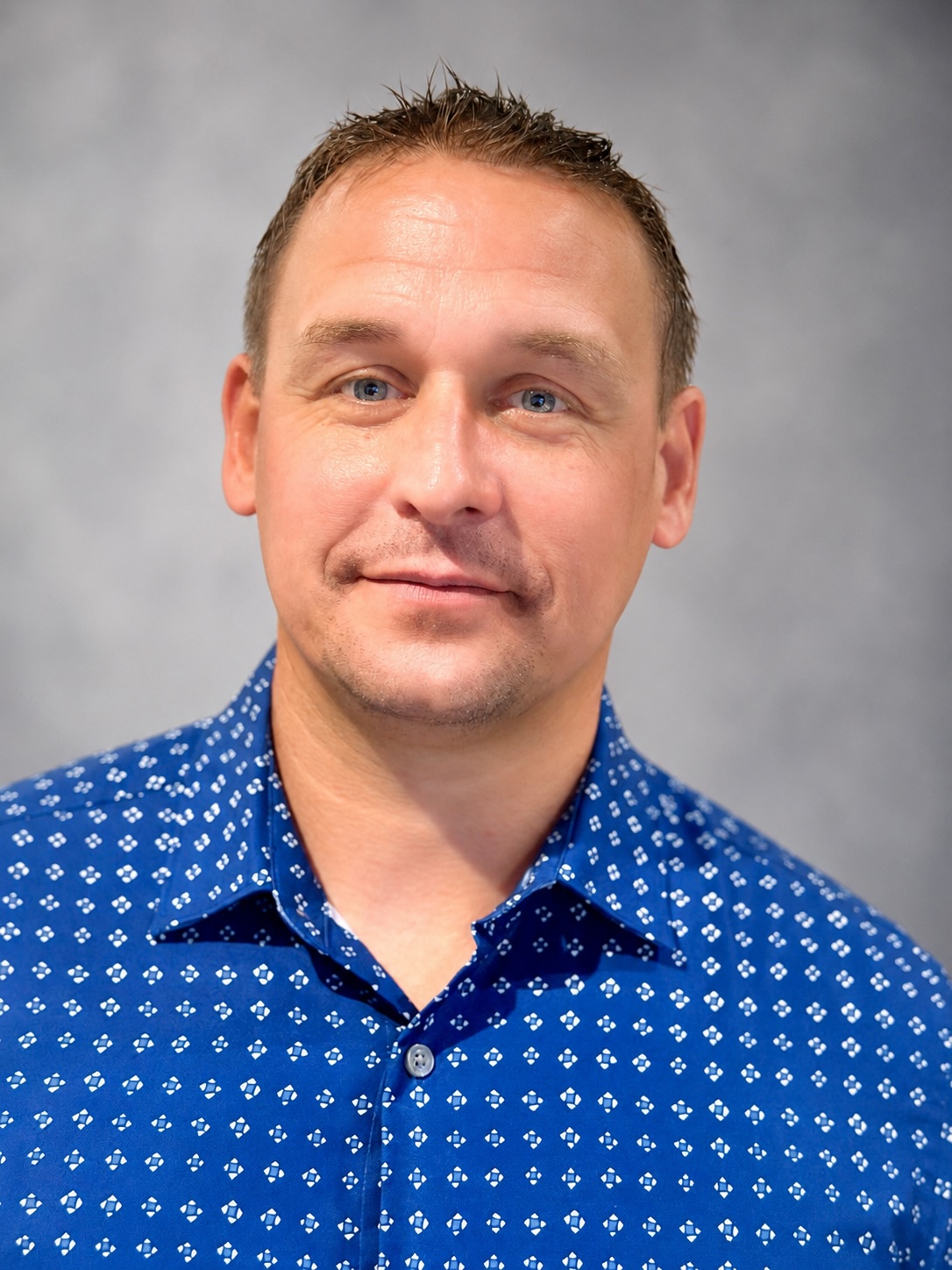 Portrait of Erin Roush, a man with short brown hair and blue eyes, wearing a blue patterned shirt.