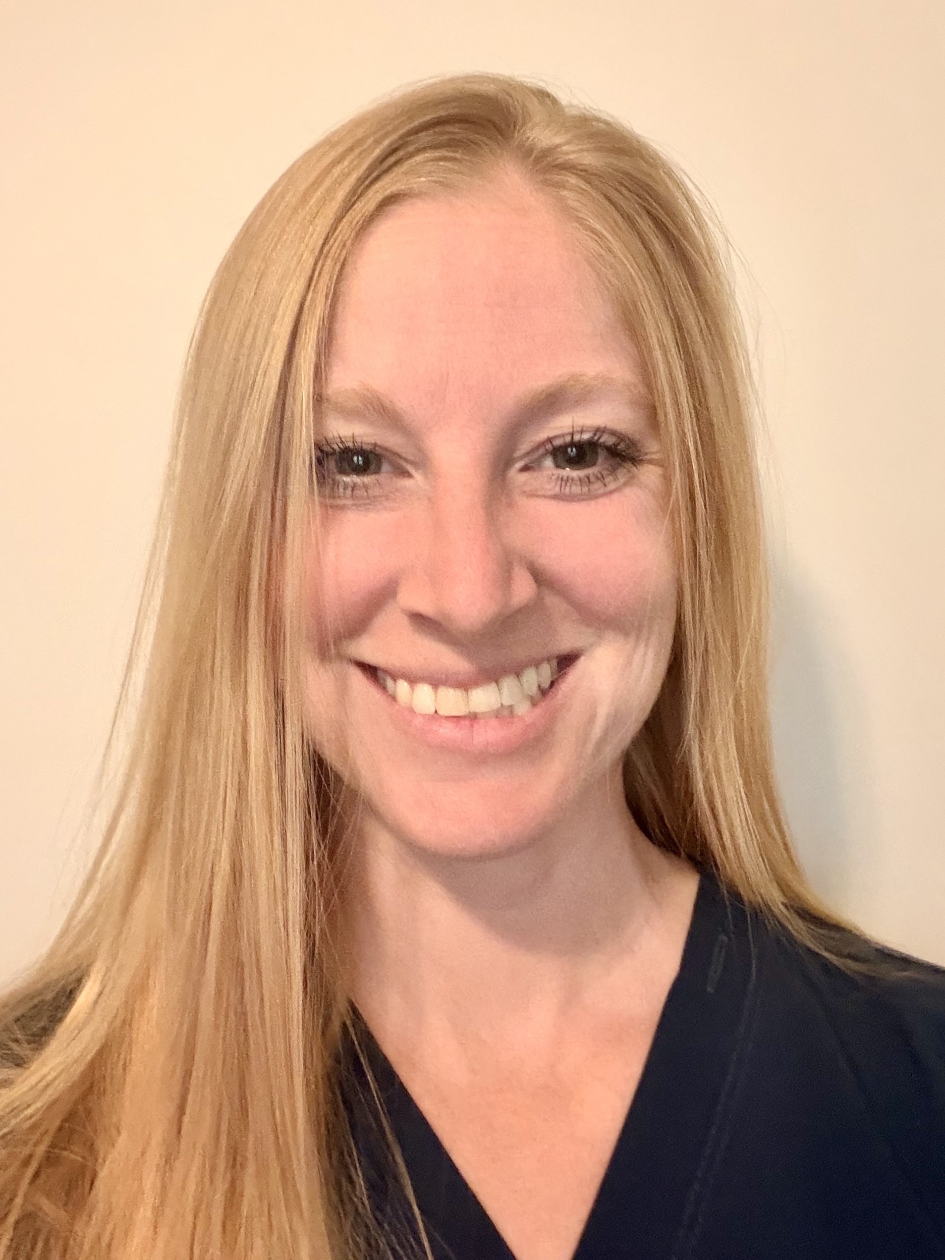 Lydia Grover, a blonde woman with a warm smile, wearing a dark blue scrub top.